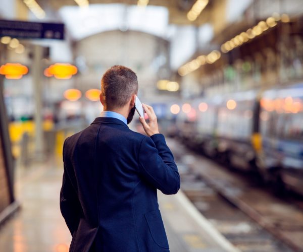Hipster businessman with smartphone, making a phone call, waiting at the train station platform, back view, rear, viewpoint