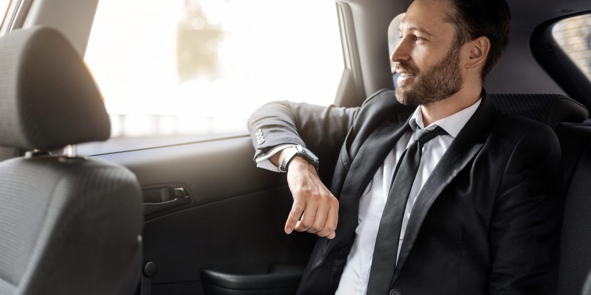 Successful businessman looking through window and smiling, going by car to meeting, thinking about business opportunities. Side view of handsome man sitting in luxury car, panorama with copy space
