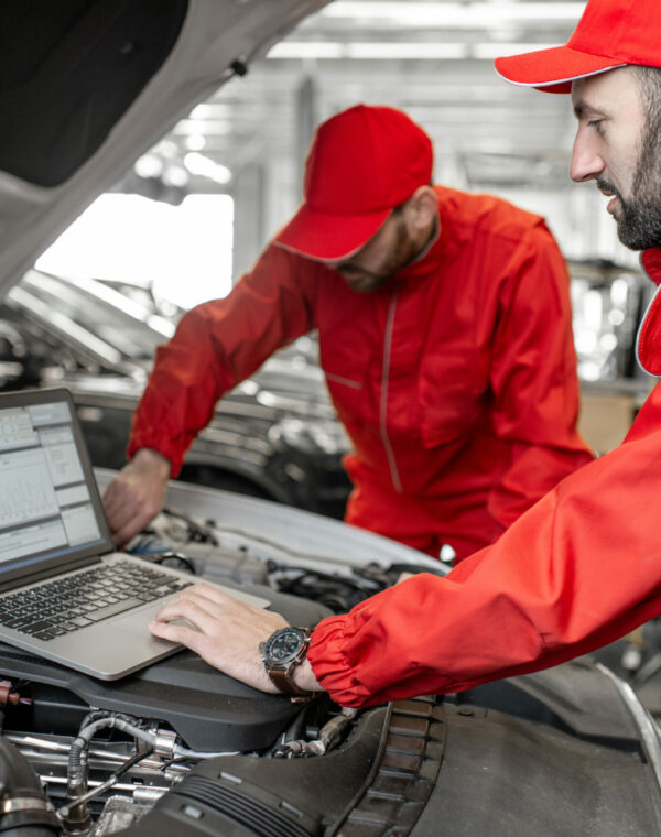 Two auto mechanics in red uniform doing engine diagnostics with computer in the car service