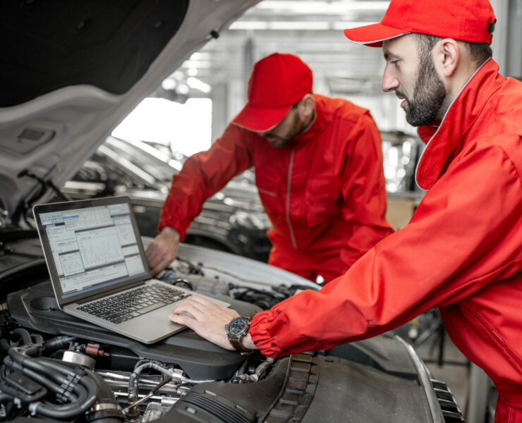 Two auto mechanics in red uniform doing engine diagnostics with computer in the car service