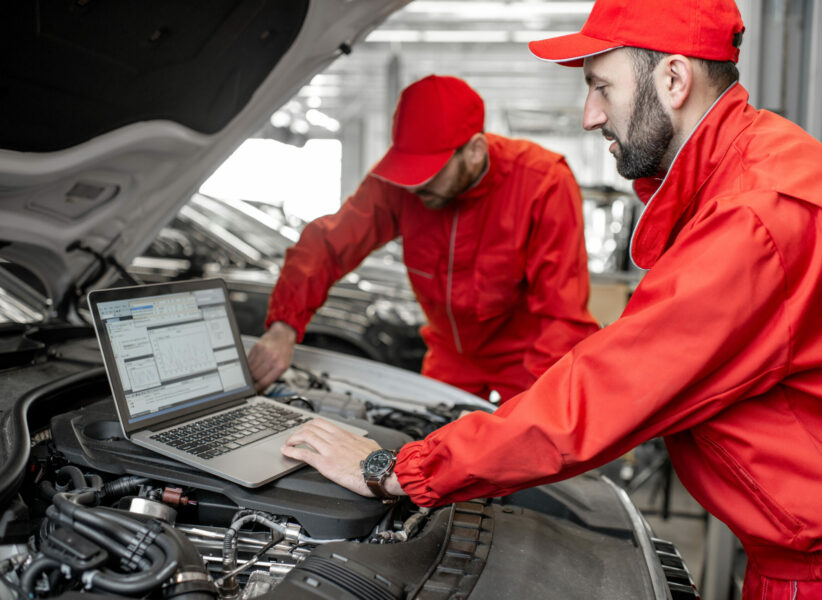 Two auto mechanics in red uniform doing engine diagnostics with computer in the car service